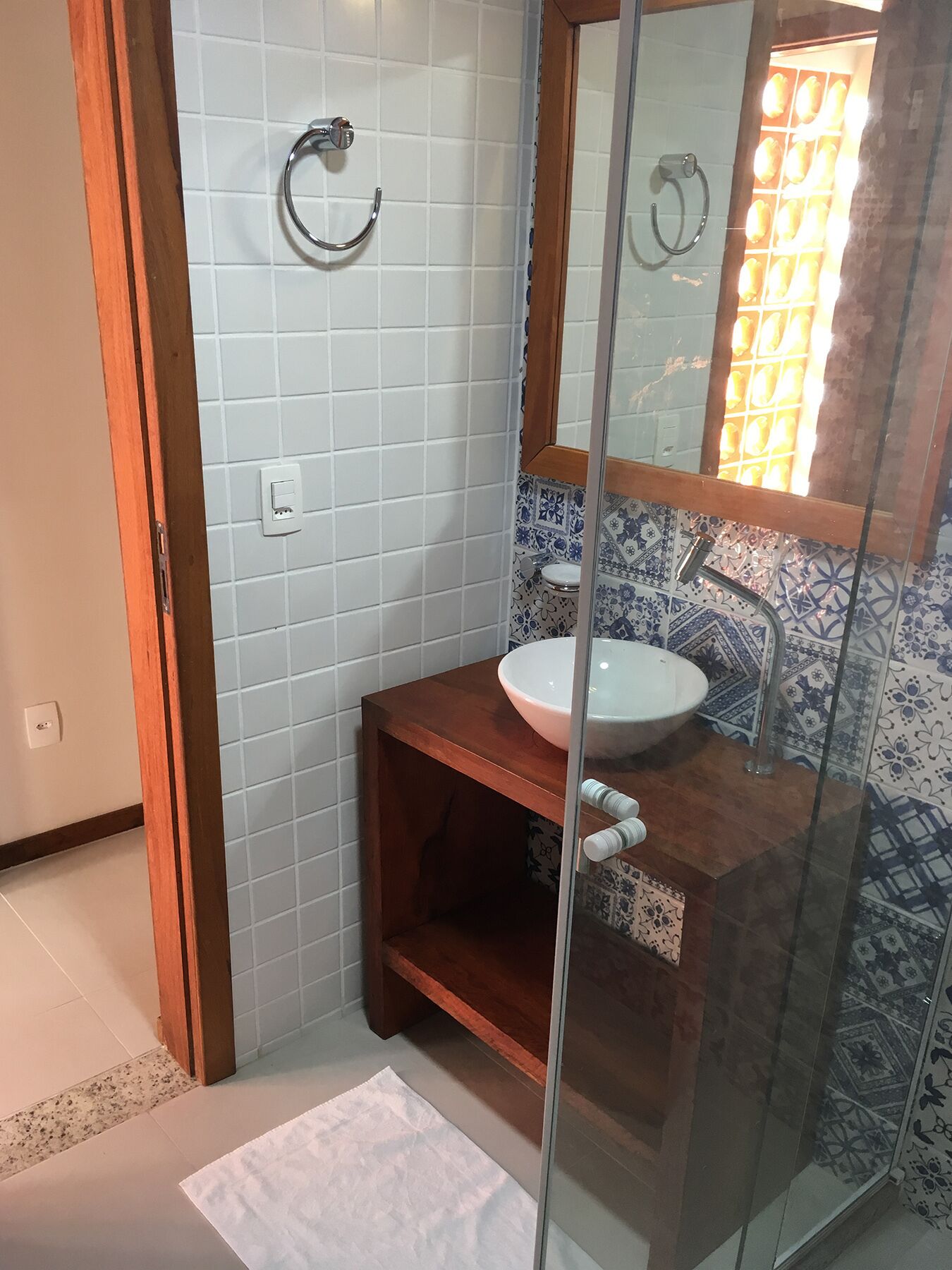 Ein Badezimmer mit einem weißen Waschbecken, einer Holzanrichte, einer Duschkabine aus Glas und einer gefliesten Wand mit floralem Muster.
