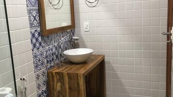 Un baño con tocador de madera, lavamanos blanco, espejo y zona de ducha.