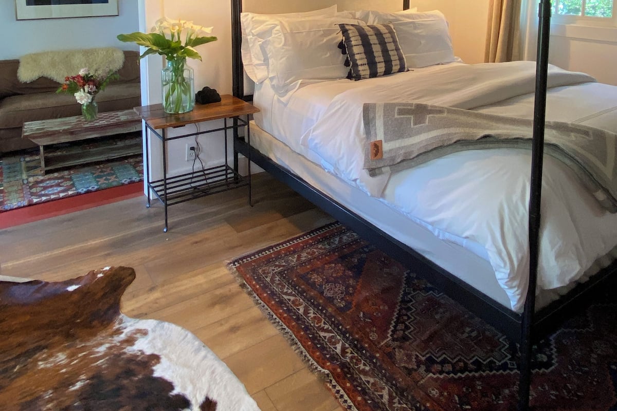 A bedroom with a four-poster bed, a bedside table with a lamp, a wooden headboard, a window with curtains, a rug, and a cowhide rug on the floor.