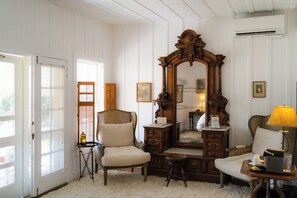 A room with a large mirror, a chair, a table with a lamp, and a small stool.
