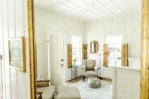 A room with a white door, a chair, a round ottoman, a table with a vase, and a mirror on the wall.