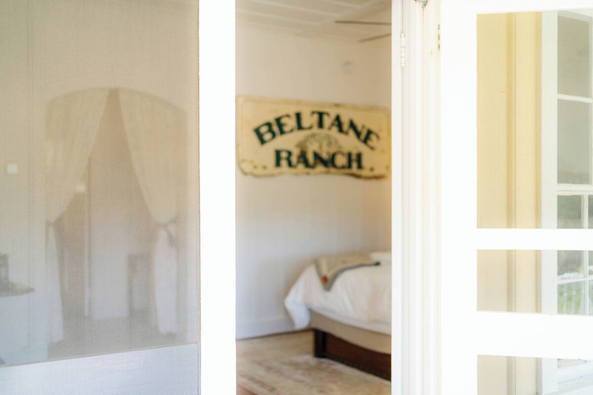 A room with a bed, a wall sign reading "BELTANE RANCH," and a window with blinds.