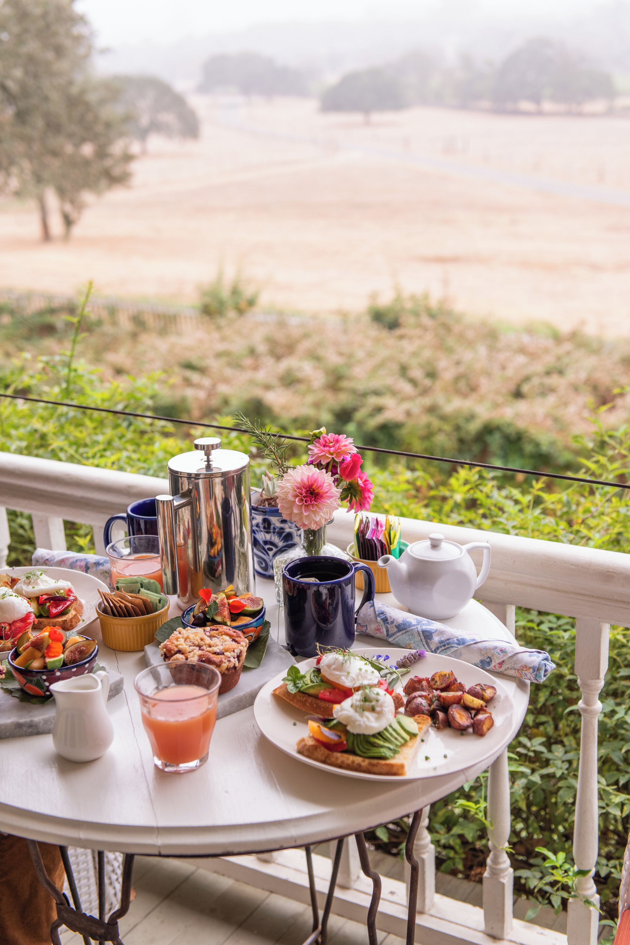 A table set with breakfast items, a pitcher, and a teapot on a balcony overlooking a landscape.