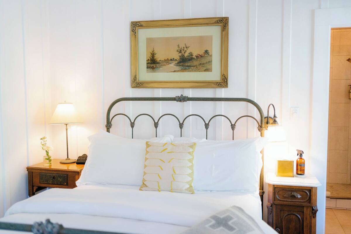 A neatly made bed with white linens, a decorative pillow, and a nightstand with a lamp and a small vase of flowers.