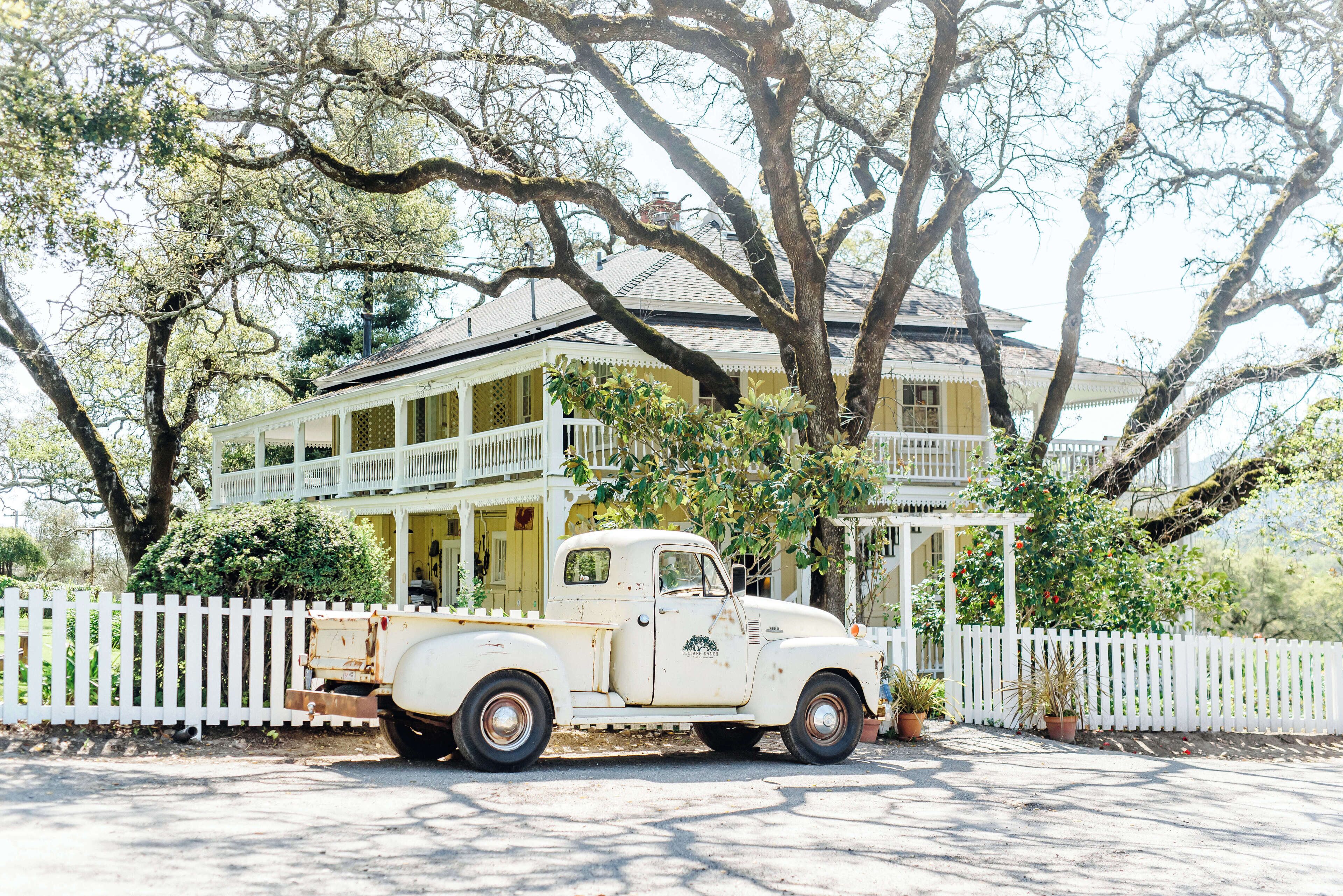 A vintage white truck parked in front of a large, yellow house with a white picket fence and a balcony.