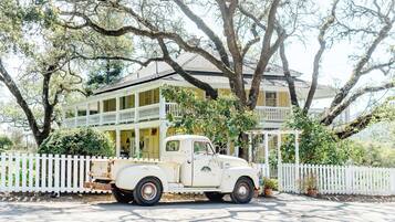A vintage white truck parked in front of a large, yellow house with a white picket fence and a balcony.