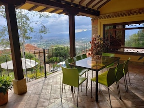 A covered patio with a table and chairs, a view of the countryside, and a balcony railing.
