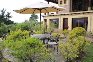 A patio with a round table and chairs, surrounded by greenery and a large umbrella.