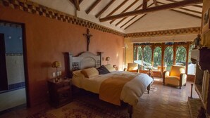 A bedroom with a large bed, a nightstand, a chair, and a window with a view of trees. - Hotel La Primavera Suites & Casas Campestres (Villa de Leyva)