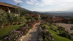 A stone pathway leading to a house with a tiled roof, surrounded by lush greenery and flowering plants. - Hotel La Primavera Suites & Casas Campestres (Villa de Leyva)