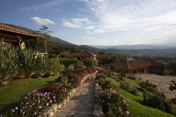A stone pathway leading to a house with a tiled roof, surrounded by lush greenery and flowering plants.