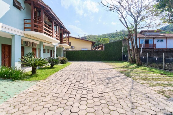 A paved courtyard with a hexagonal tile pattern, surrounded by buildings with balconies and a green hedge.