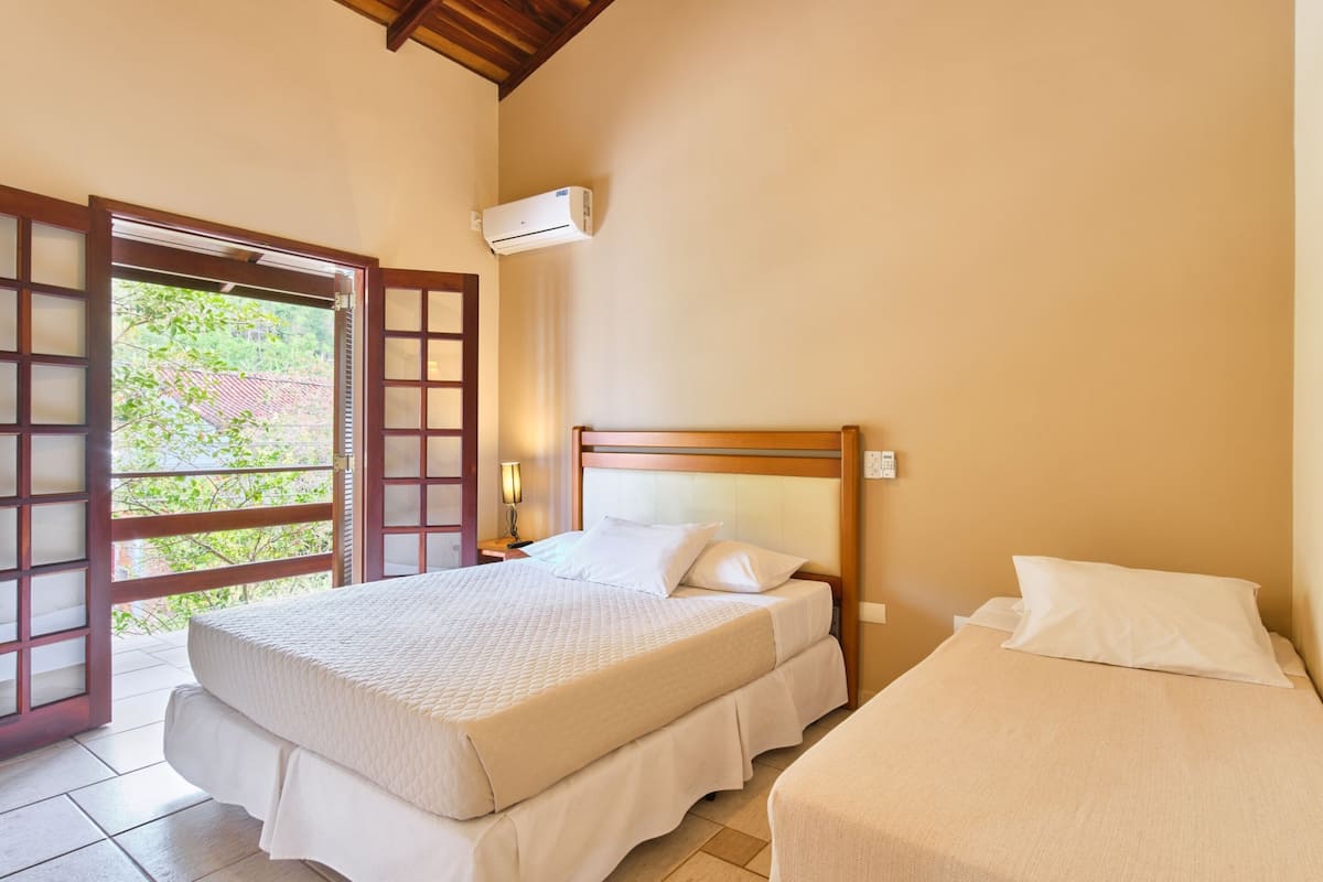 A bedroom with two beds, a wooden headboard, a window with a view of greenery, and an air conditioning unit.