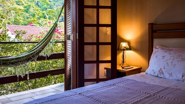 Un dormitorio con cama, mesita de noche, lámpara, ventana con vistas a un edificio de tejados rojos y vegetación, y una hamaca.
