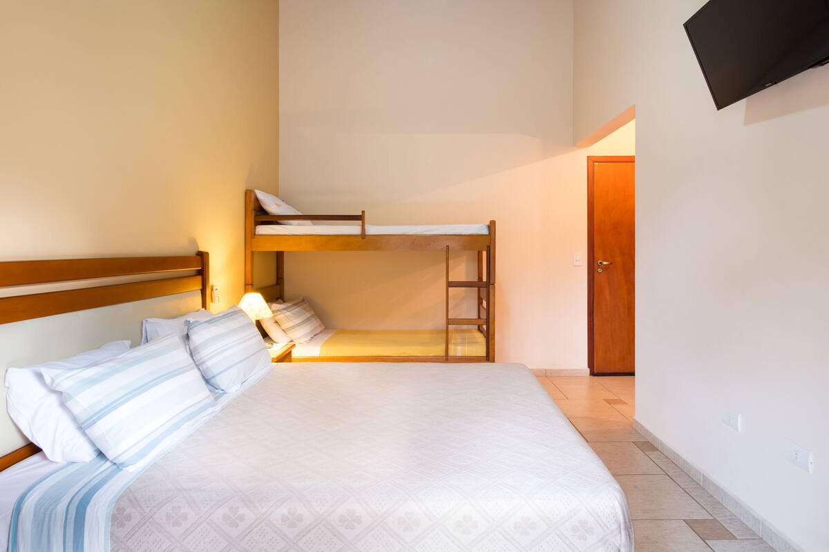 A bedroom with a bunk bed, a single bed, a wooden headboard, a bedside table with a lamp, and a TV mounted on the wall.