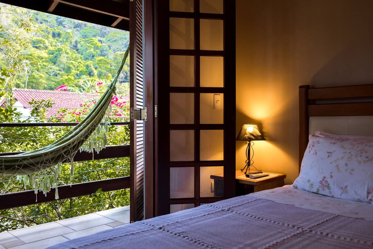 A bedroom with a bed, a nightstand, a lamp, a window with a view of greenery and a building, and a hammock.