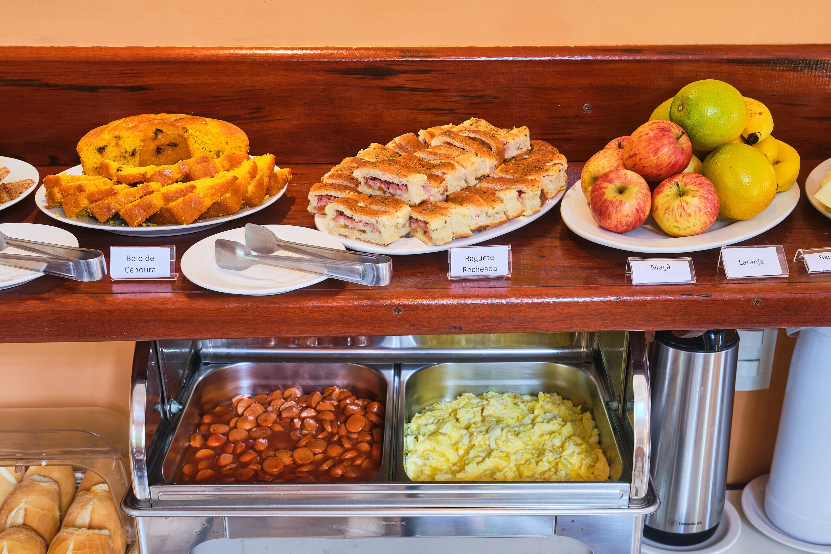 A buffet with various food items including a Bolo de Cenoura, Baguete Recheada, Maçã, and Laranja.