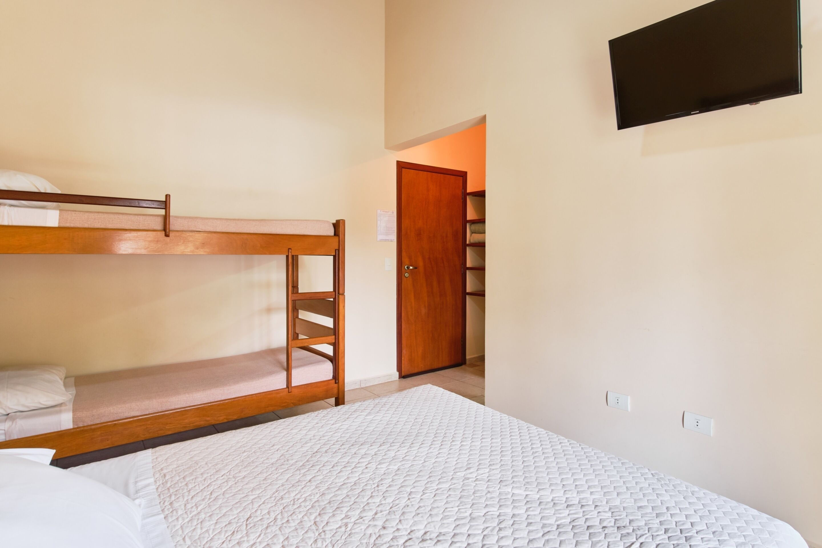 A room with bunk beds, a white quilted mattress, a wooden ladder, a door, and a television mounted on the wall.