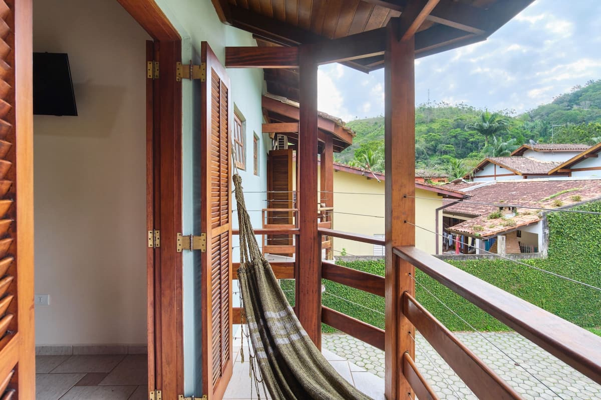 A balcony with a hammock, wooden railings, and a view of a hillside with houses.
