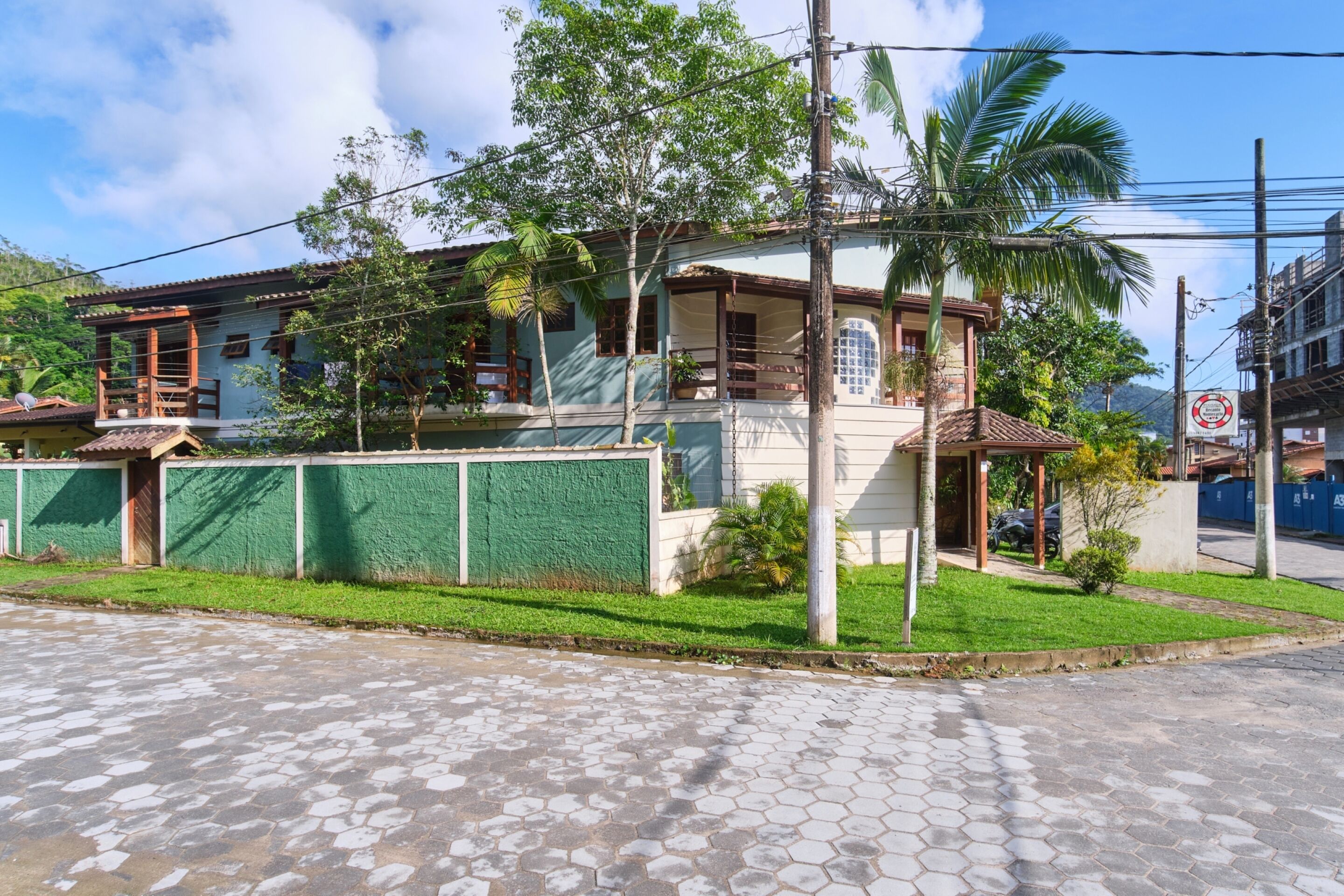 A two-story house with a balcony, surrounded by greenery and a paved street.
