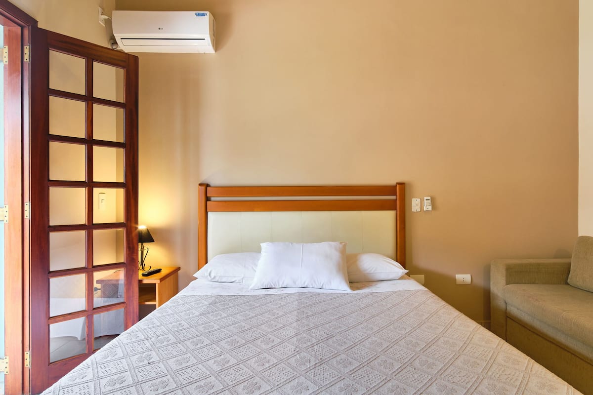 A bedroom with a wooden headboard, a bed with patterned bedding, a bedside table with a lamp, and a sofa.