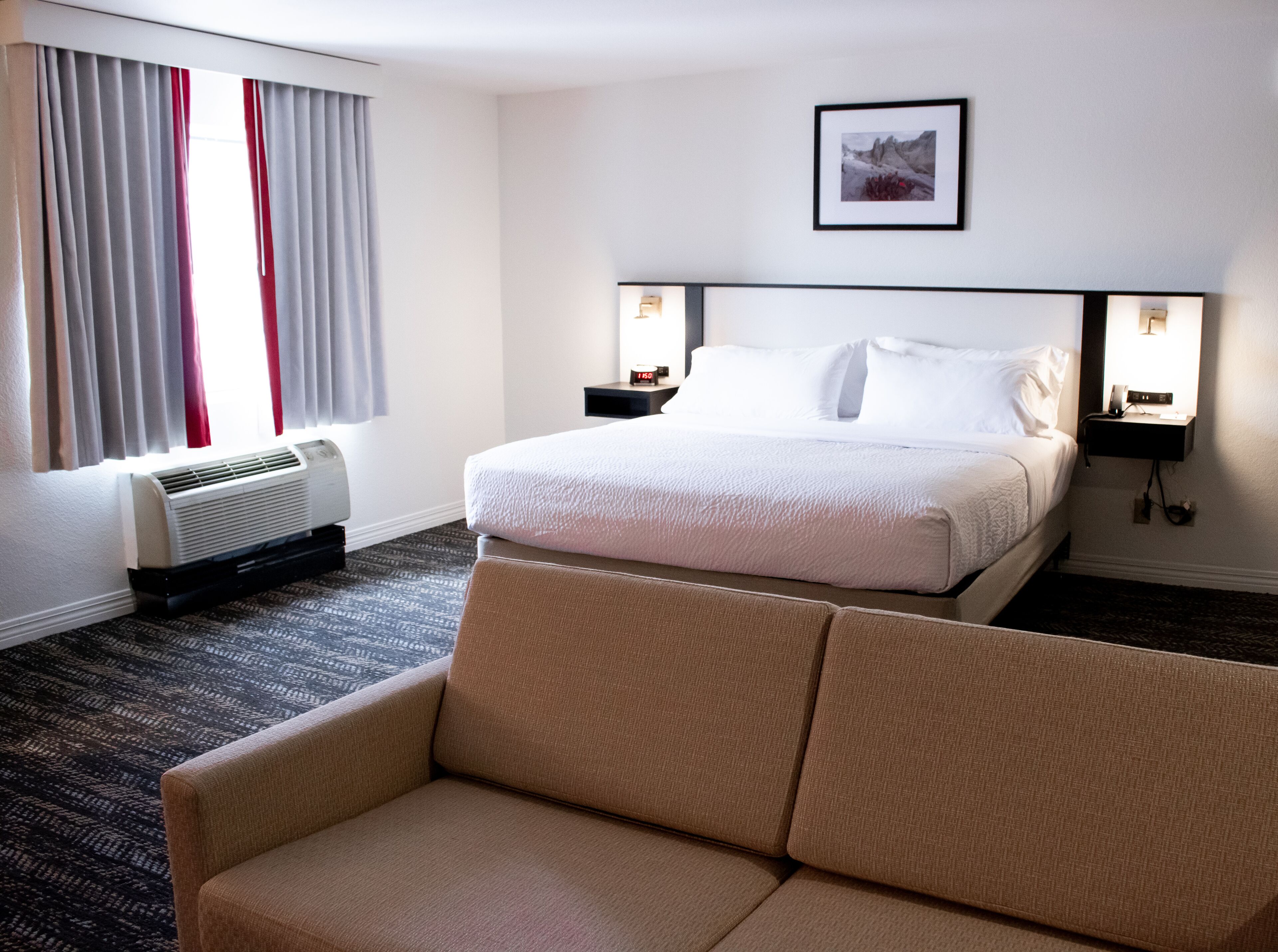 A hotel room with a large bed, a sofa, a wall-mounted picture, and a bedside table with a lamp.