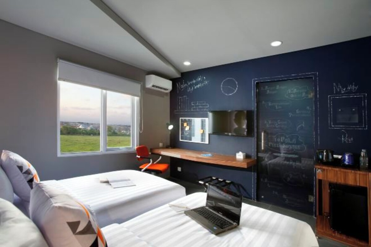 A modern hotel room with two beds, a desk, a laptop, and a large window offering a view of the outdoors.