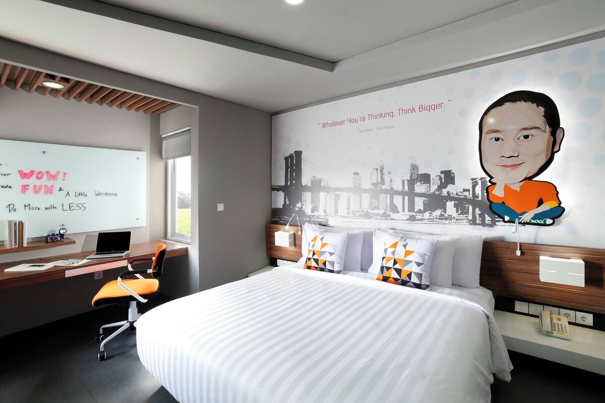 A modern hotel room with a large bed, a desk with a laptop, a whiteboard with motivational quotes, and a cityscape mural on the wall.