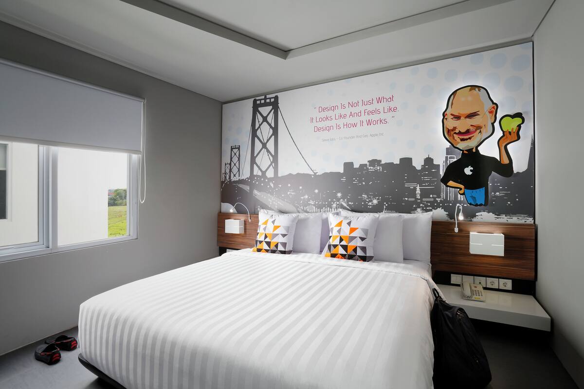 A modern hotel room with a large bed, a cityscape mural on the wall, and a view of the outdoors through a window.