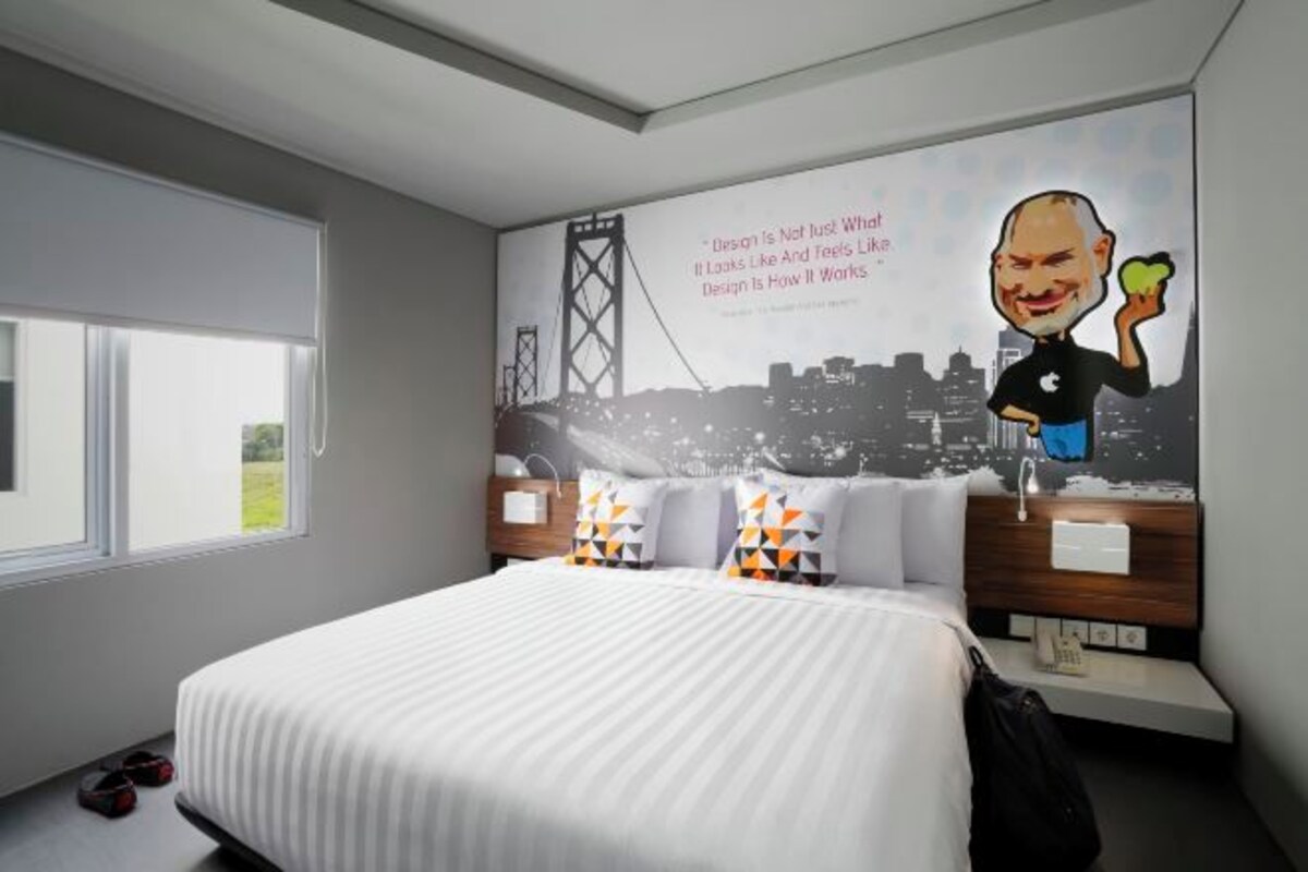 A modern hotel room with a large bed, a cityscape mural on the wall, and a view of the outdoors through the window.