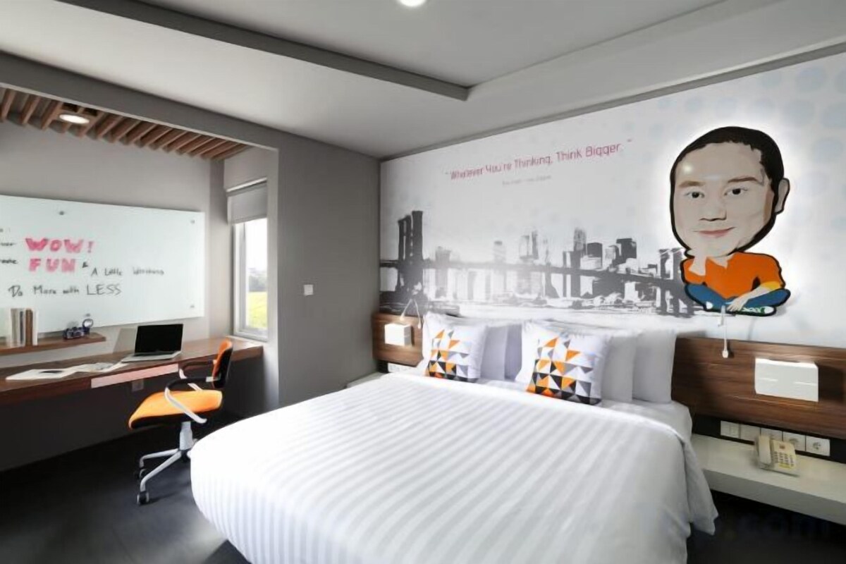 A hotel room with a bed, a desk with a laptop, a whiteboard with motivational quotes, and a cityscape mural on the wall.