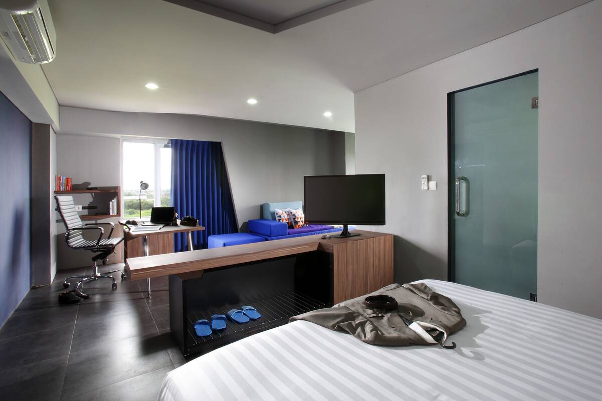 A modern hotel room with a bed, a desk with a computer, a TV, and a small seating area with a sofa and a chair.