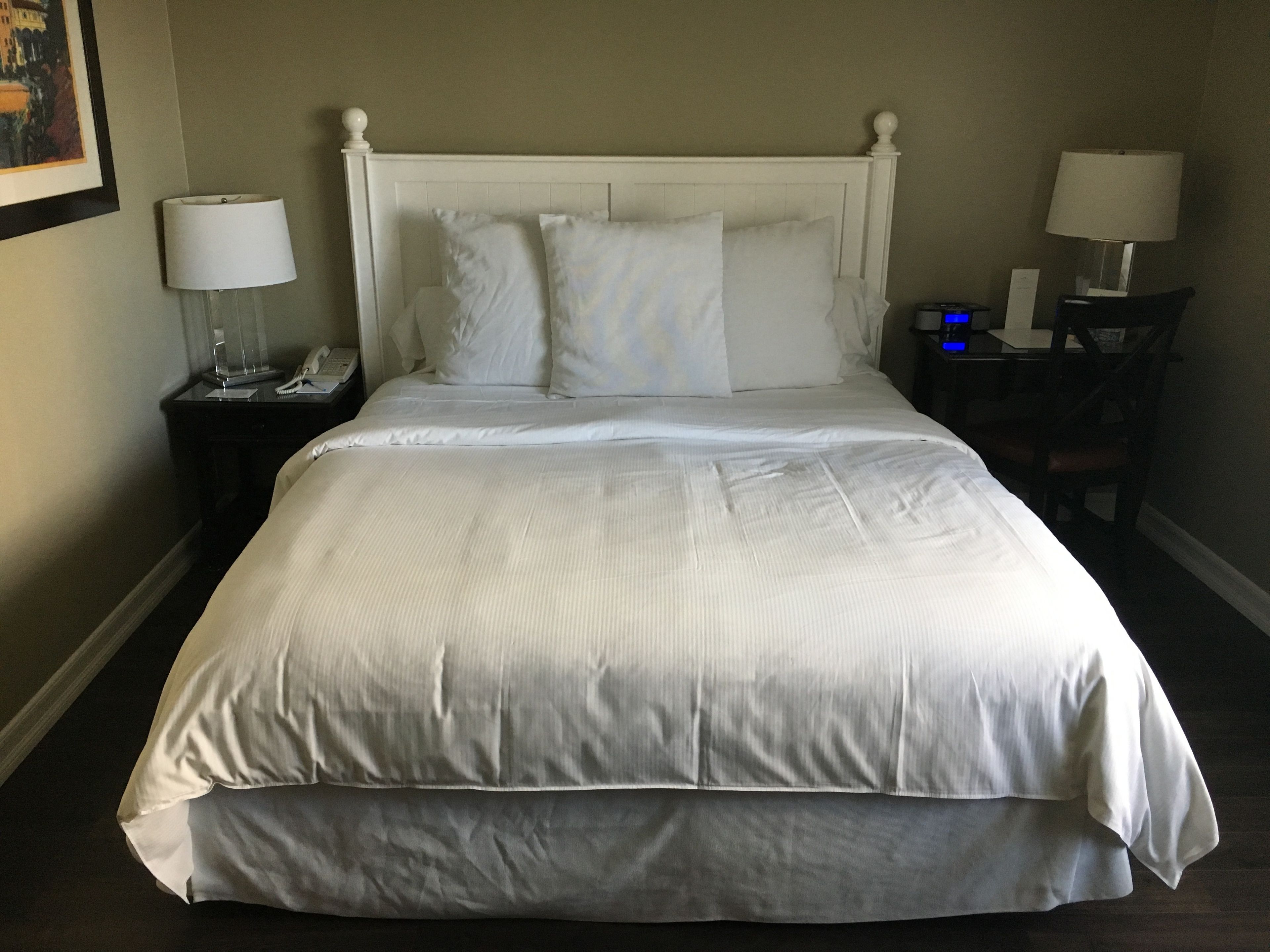 A neatly made bed with white linens, a bedside table with a lamp, and a chair with a table.