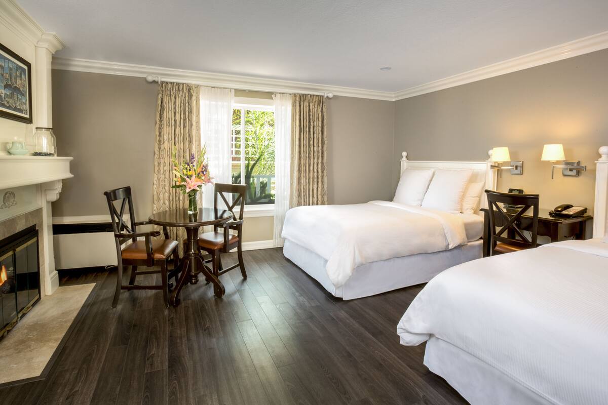 Una habitación de hotel con dos camas, una mesa de comedor con sillas, una chimenea y una ventana con cortinas.
