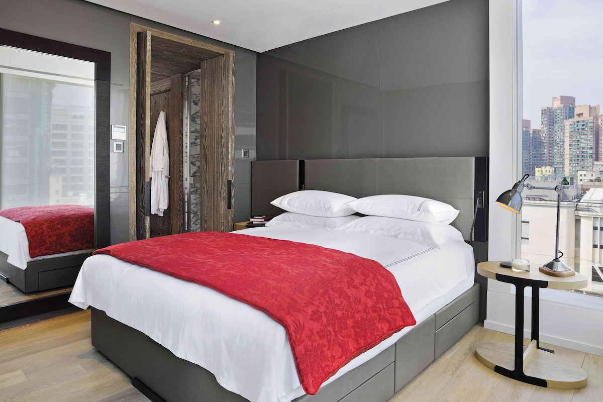 A modern hotel room with a large bed, a bedside table, a mirror, and a city view.