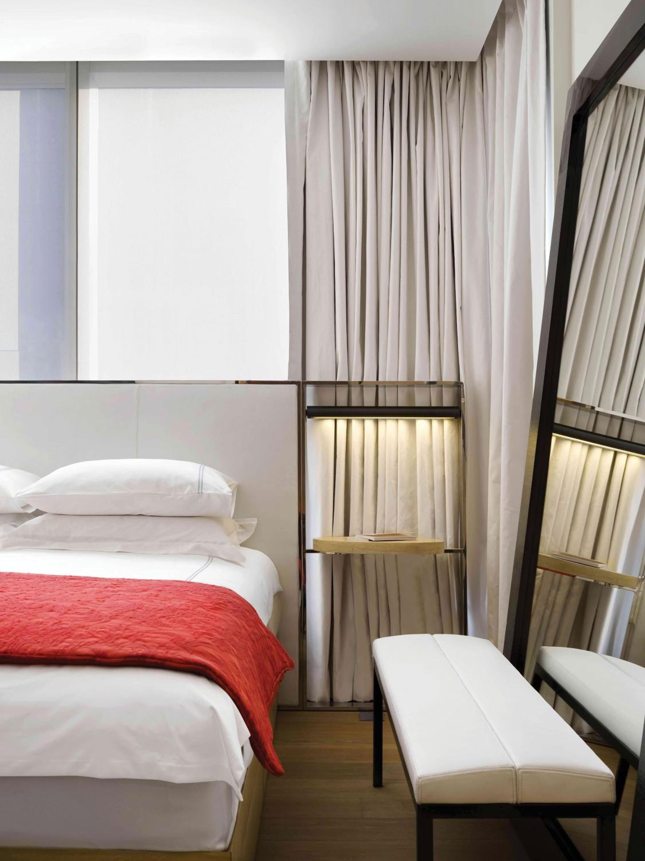 A hotel room with a bed, a bench, a bedside table, and a window with curtains.
