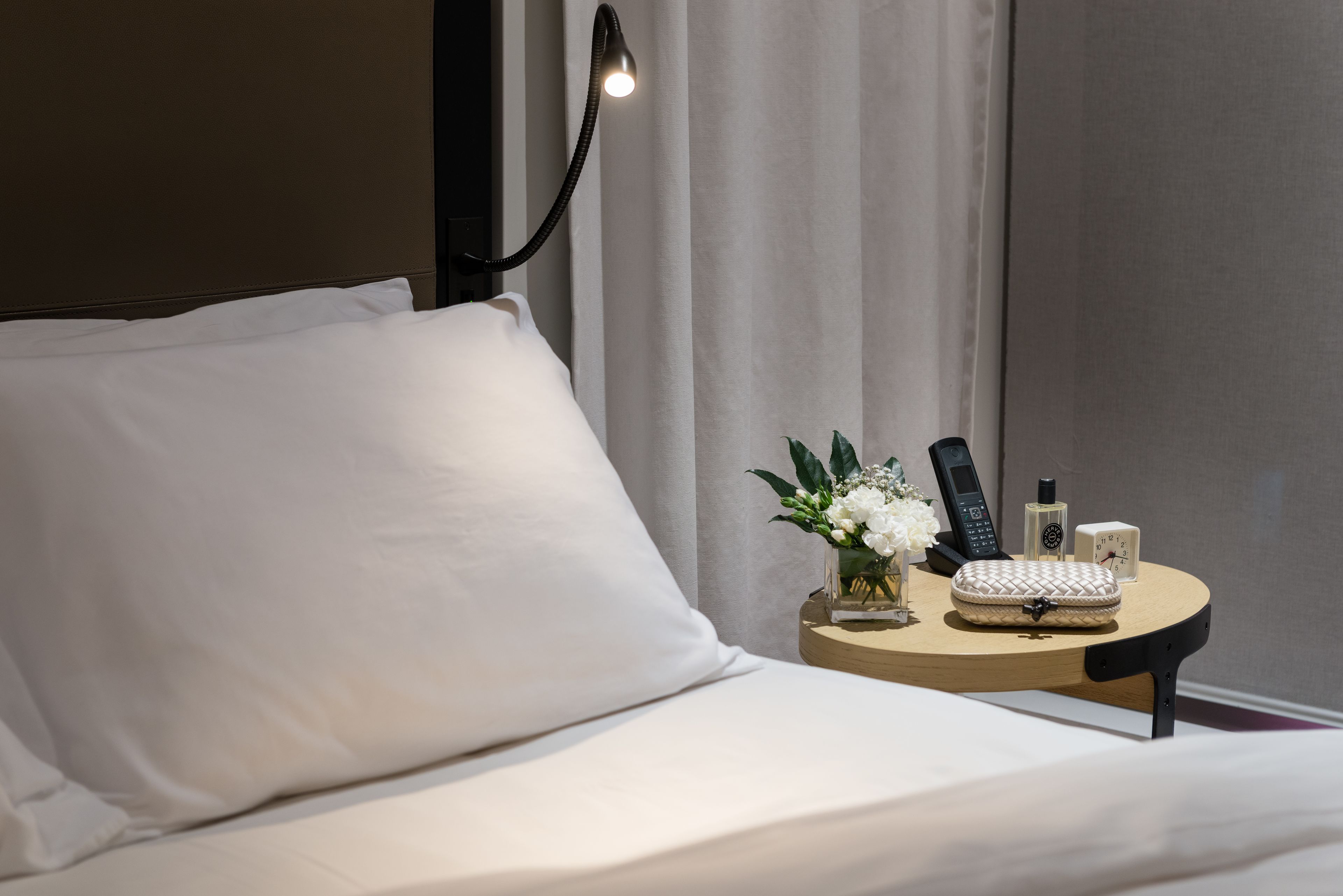 Een net opgemaakt bed met witte lakens, een nachtkastje met een lamp, een vaas met bloemen, een telefoon, een fles en een wekker.