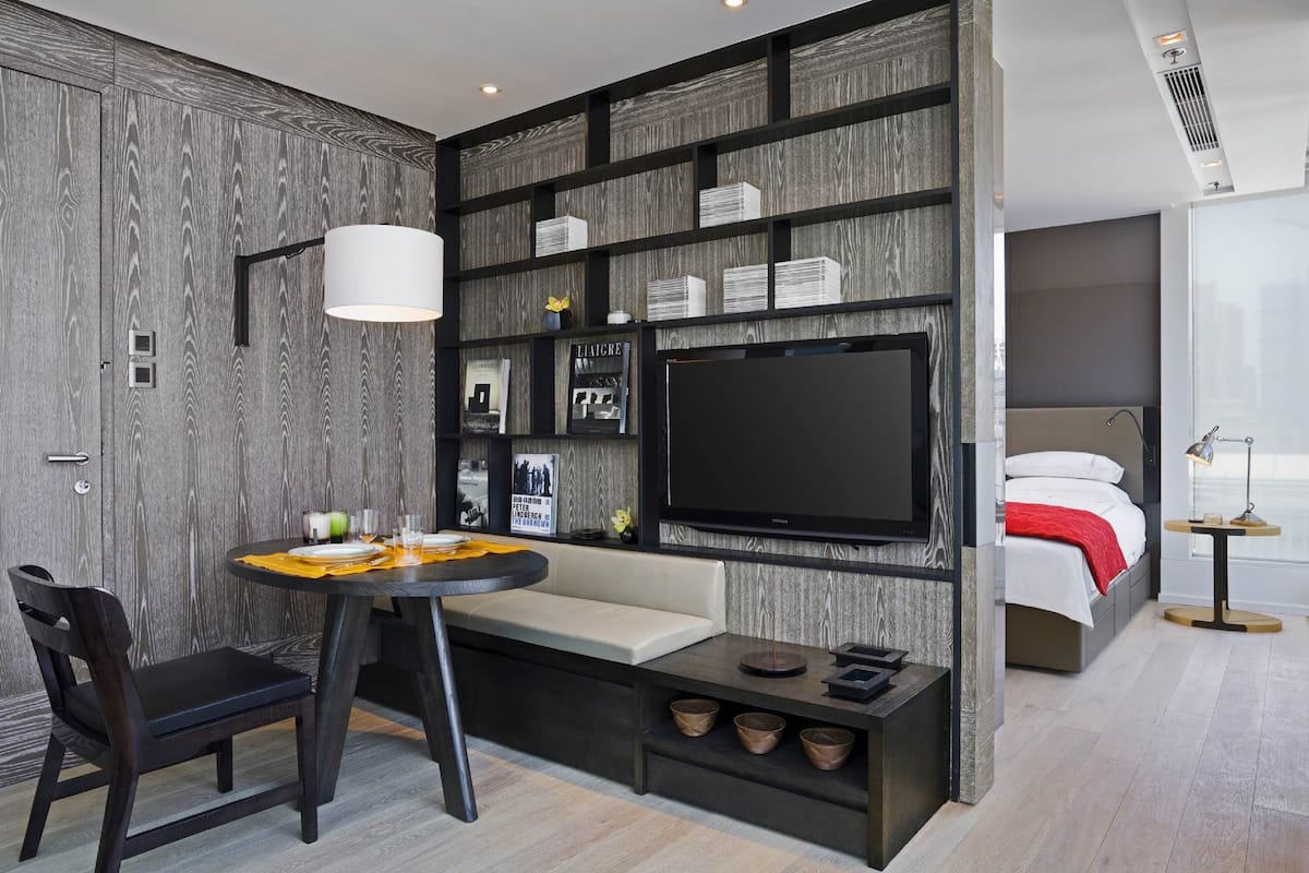 Une chambre d’hôtel moderne dotée d’un grand téléviseur à écran plat, d’un coin repas avec une table ronde et des chaises, d’une banquette intégrée et d’une table de chevet avec une lampe.