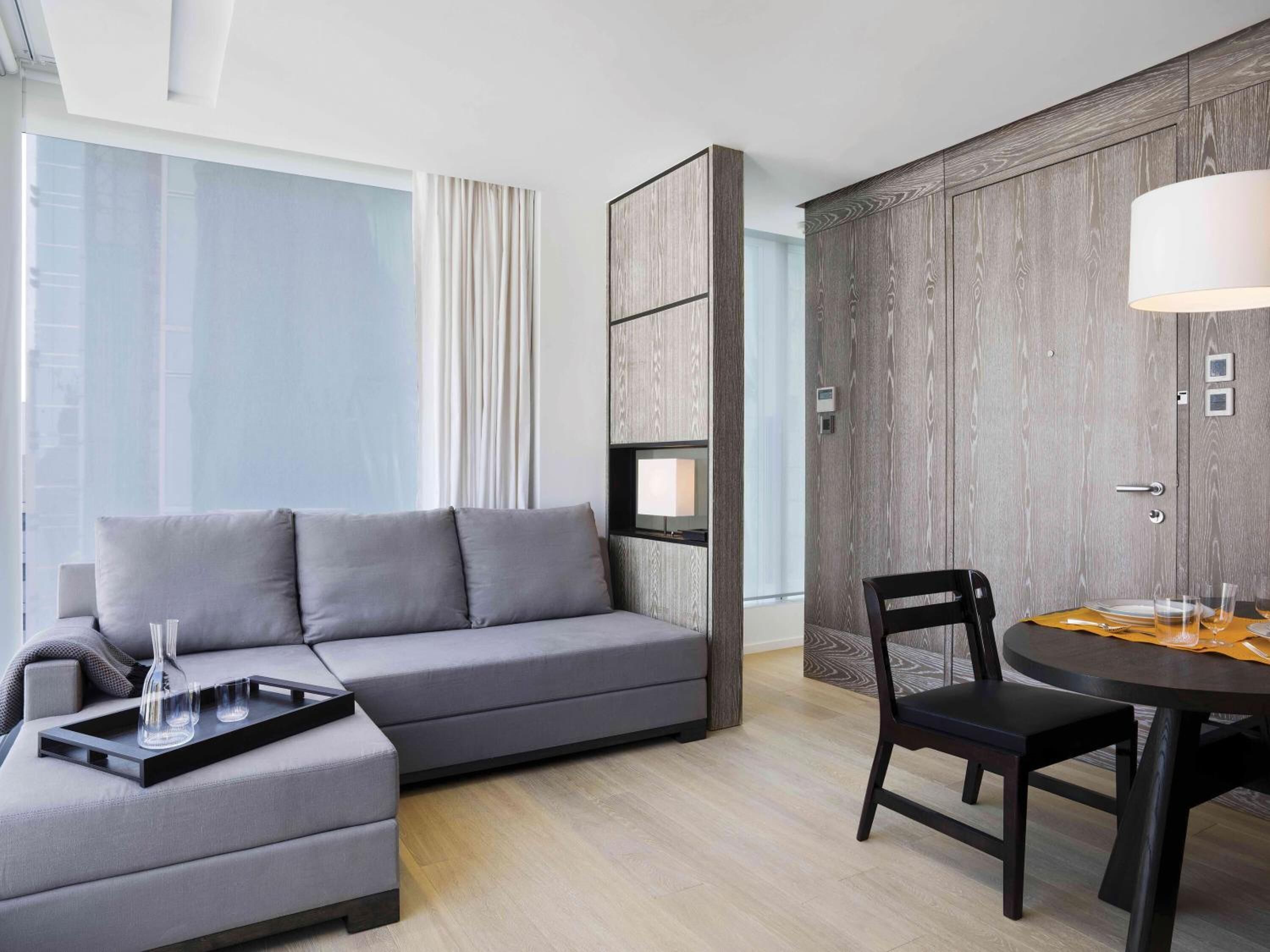 Une chambre d’hôtel moderne avec un canapé gris, une table à manger avec des chaises, une armoire en bois et une grande fenêtre avec des rideaux.