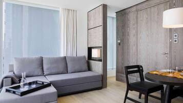 Une chambre d’hôtel moderne avec un canapé gris, une table à manger avec des chaises, une armoire en bois et une grande fenêtre avec des rideaux.