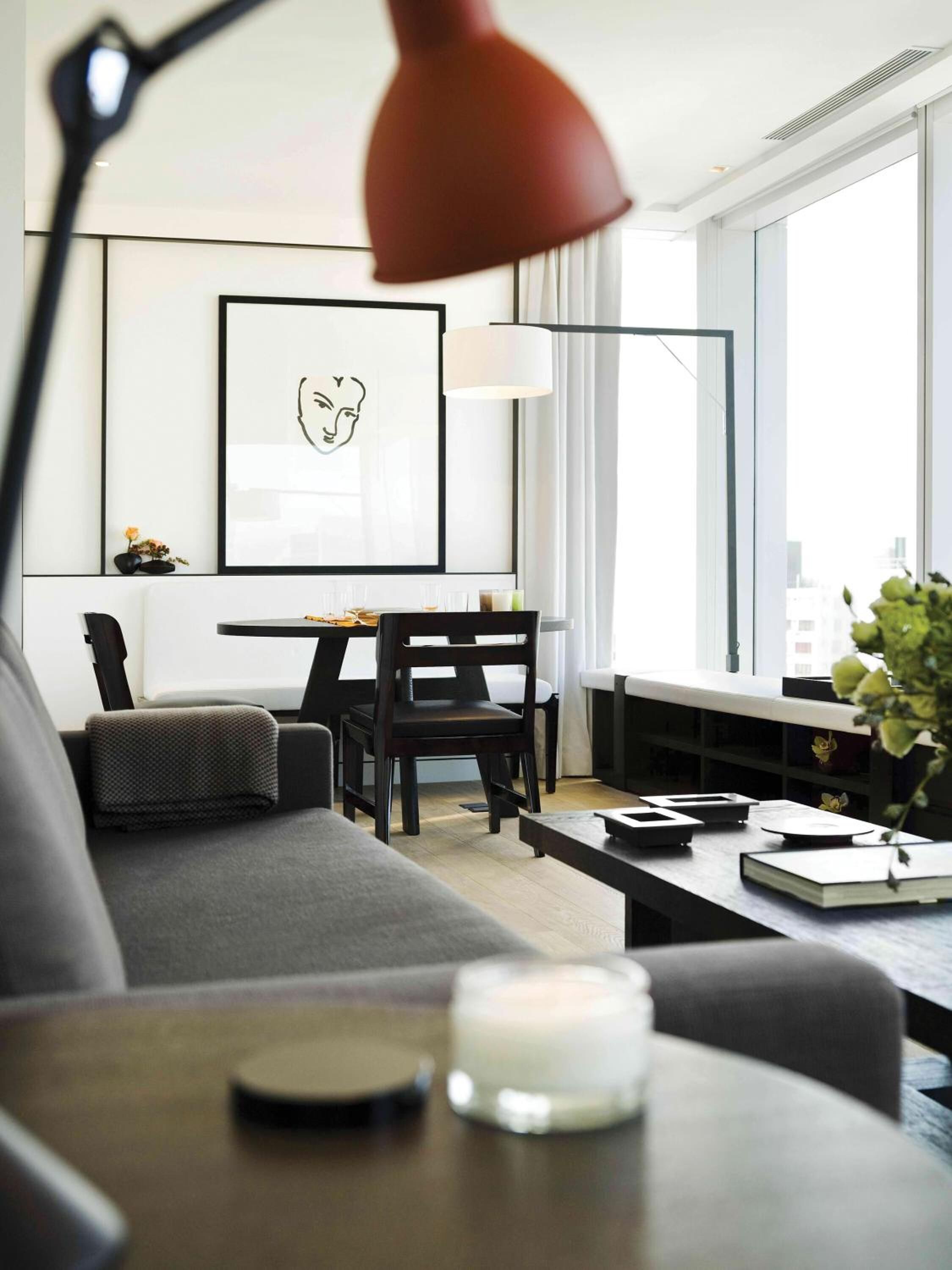 A modern living room with a sofa, a dining table, and a desk. There is a framed artwork on the wall, a lamp, and a window providing natural light.