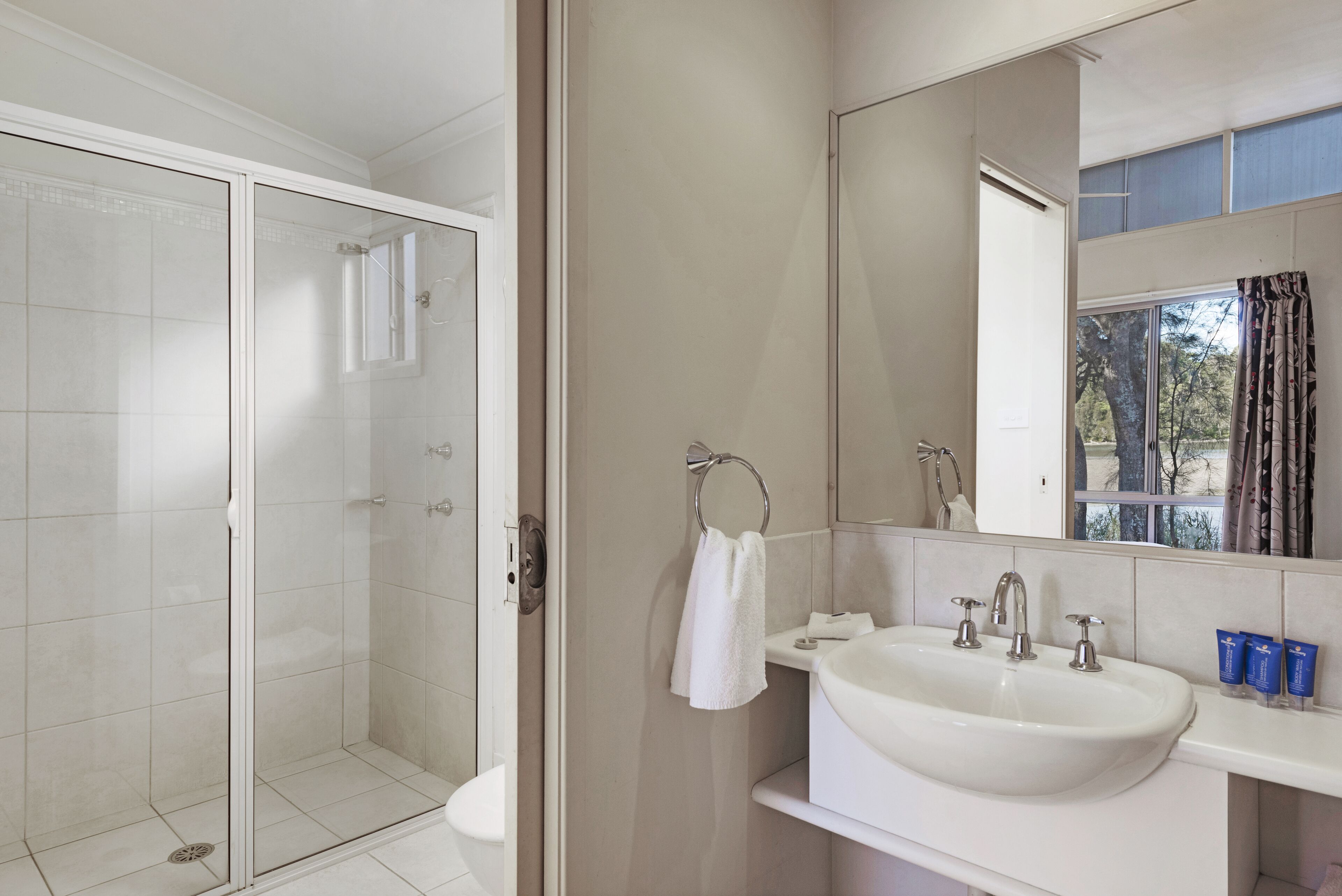 A bathroom with a glass shower enclosure, a sink with a mirror, and a window with curtains.