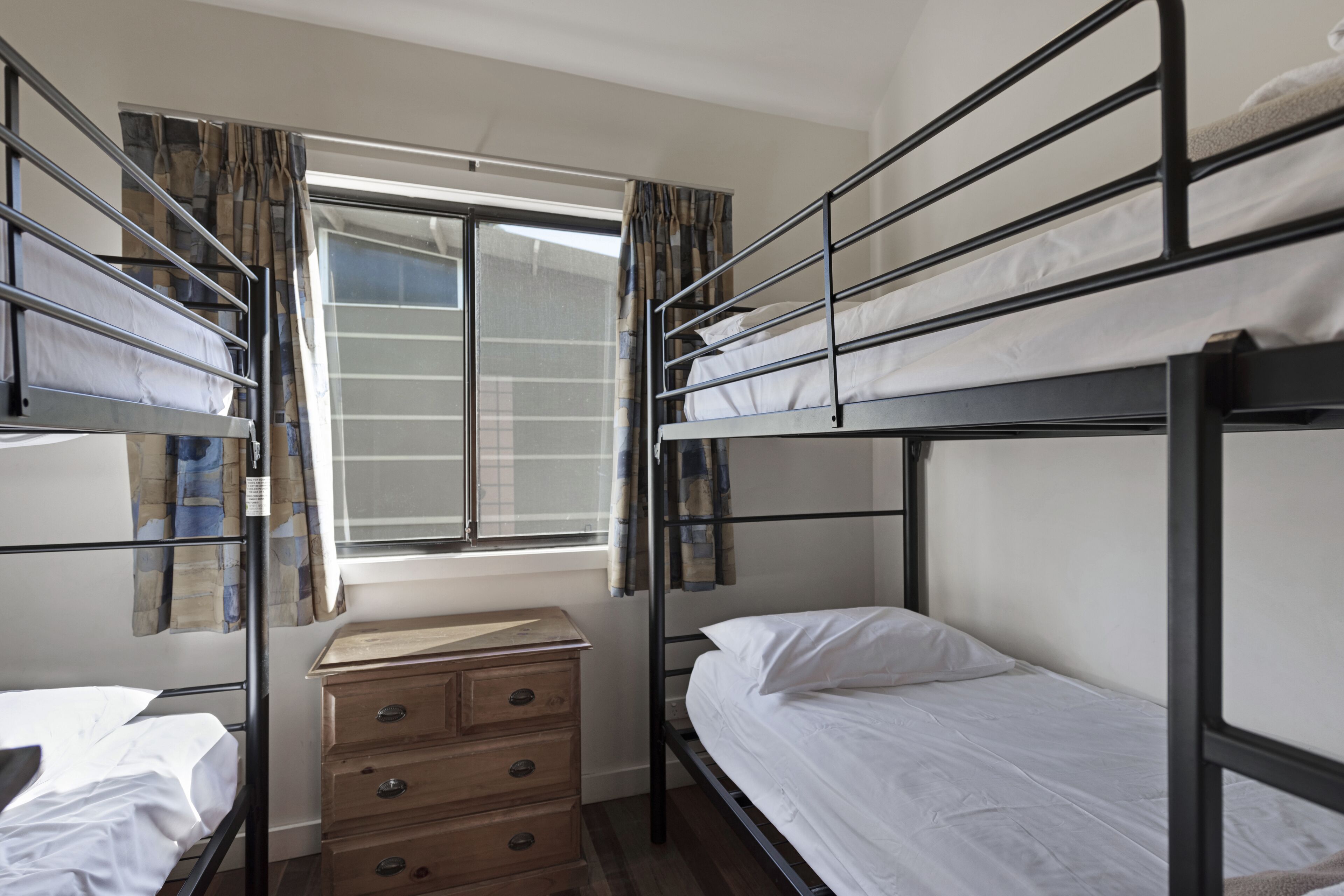A dormitory room with bunk beds, a window with curtains, a wooden dresser, and a small desk.