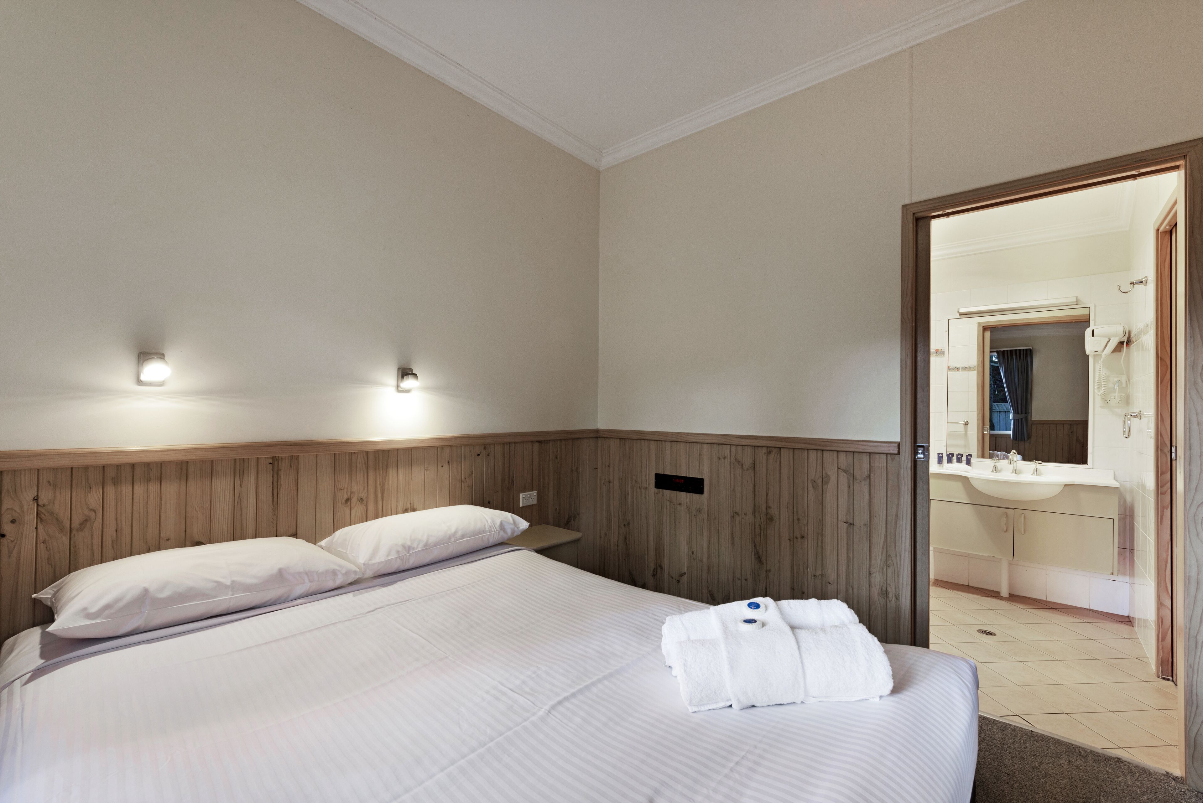 Ein Hotelzimmer mit einem Holzkopfteil, zwei Betten mit weißer Bettwäsche, einem Badezimmer mit Waschbecken und Spiegel sowie einem Handtuch auf dem Bett.