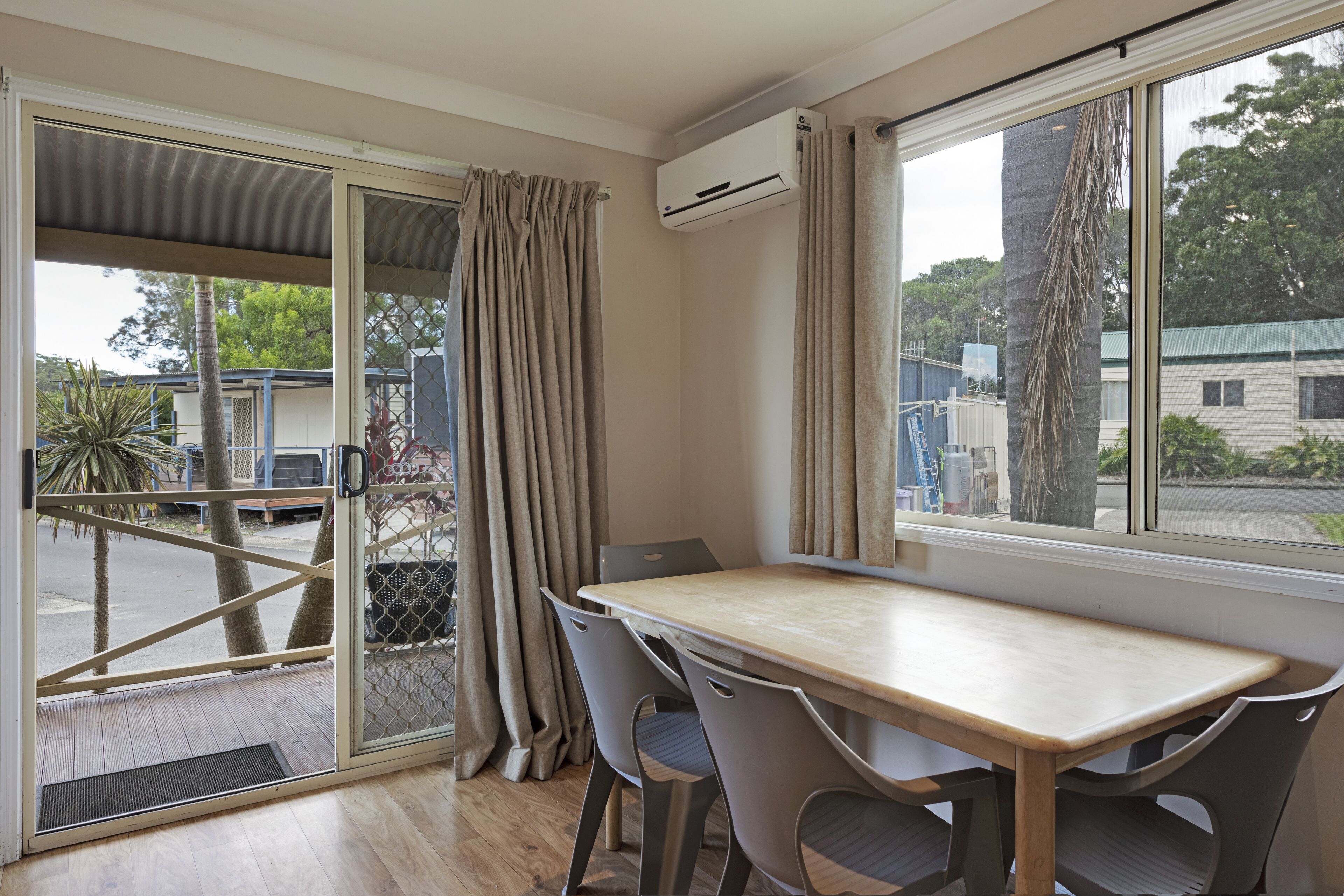 Una habitación con una mesa y sillas de madera, una puerta corredera de vidrio con cortinas, un equipo de aire acondicionado y vistas al exterior.