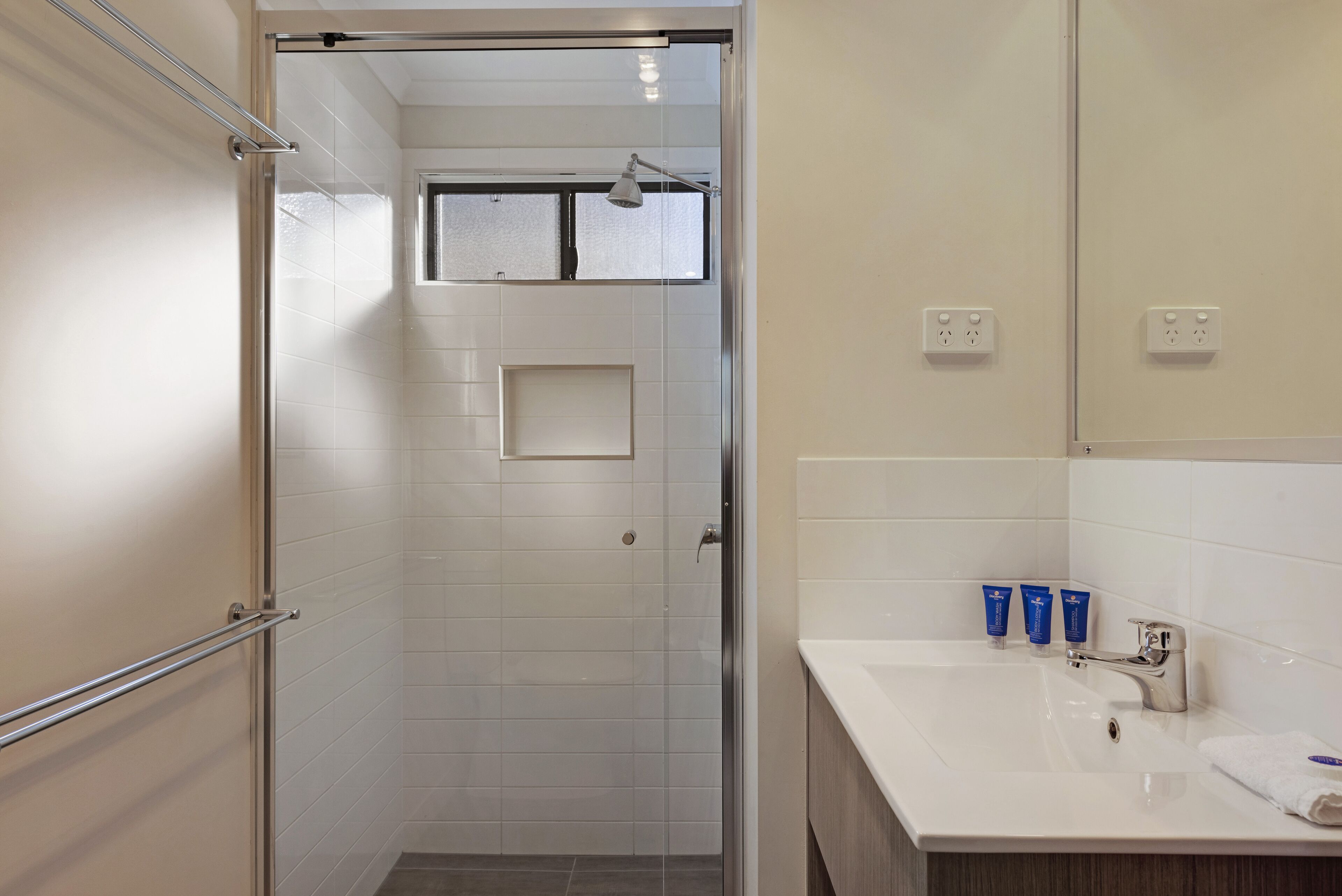 A modern bathroom with a glass shower enclosure, a white sink with a mirror, and two electrical outlets on the wall.