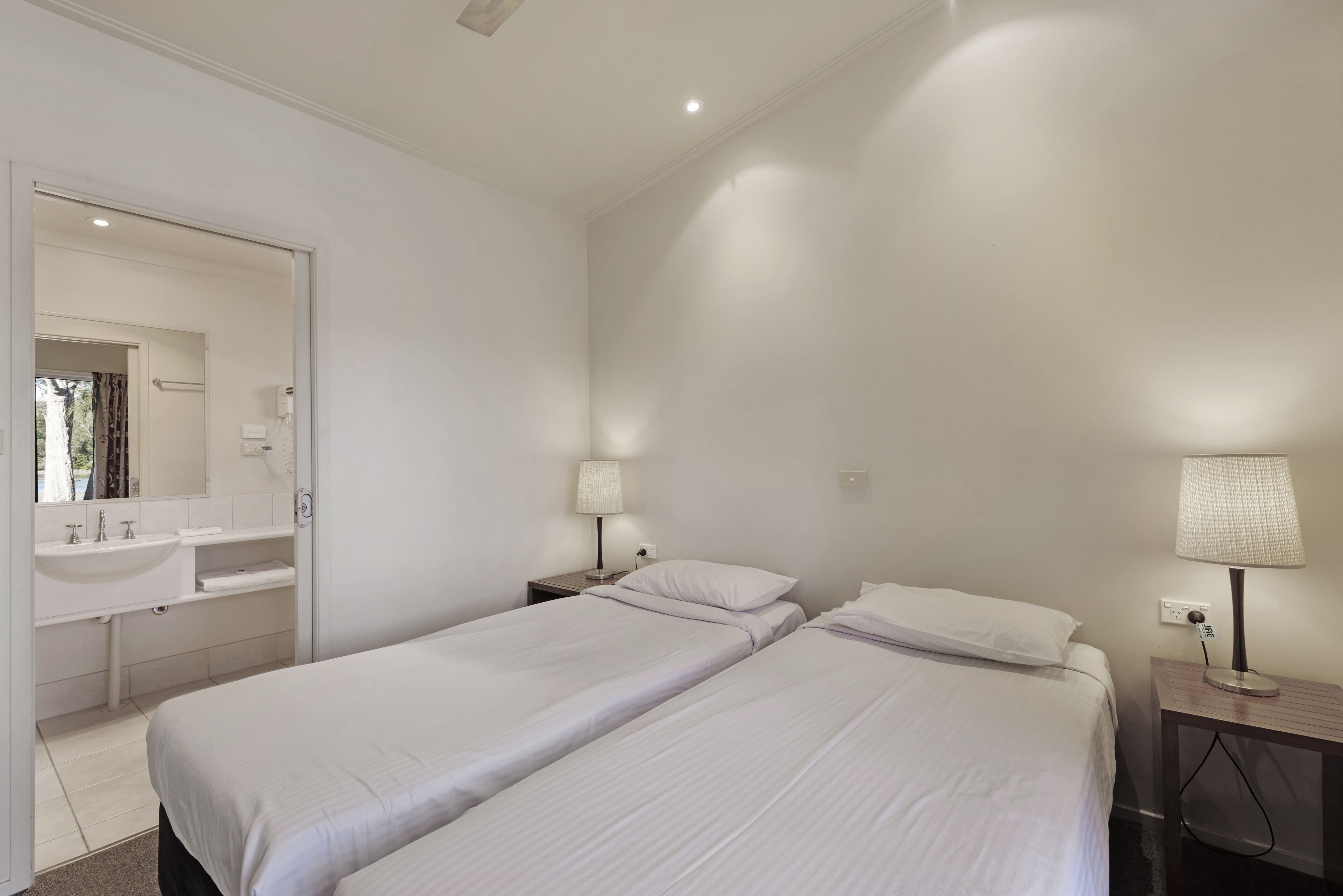 A clean, modern hotel room with two single beds, a bathroom with a sink and mirror, and a bedside table with a lamp.