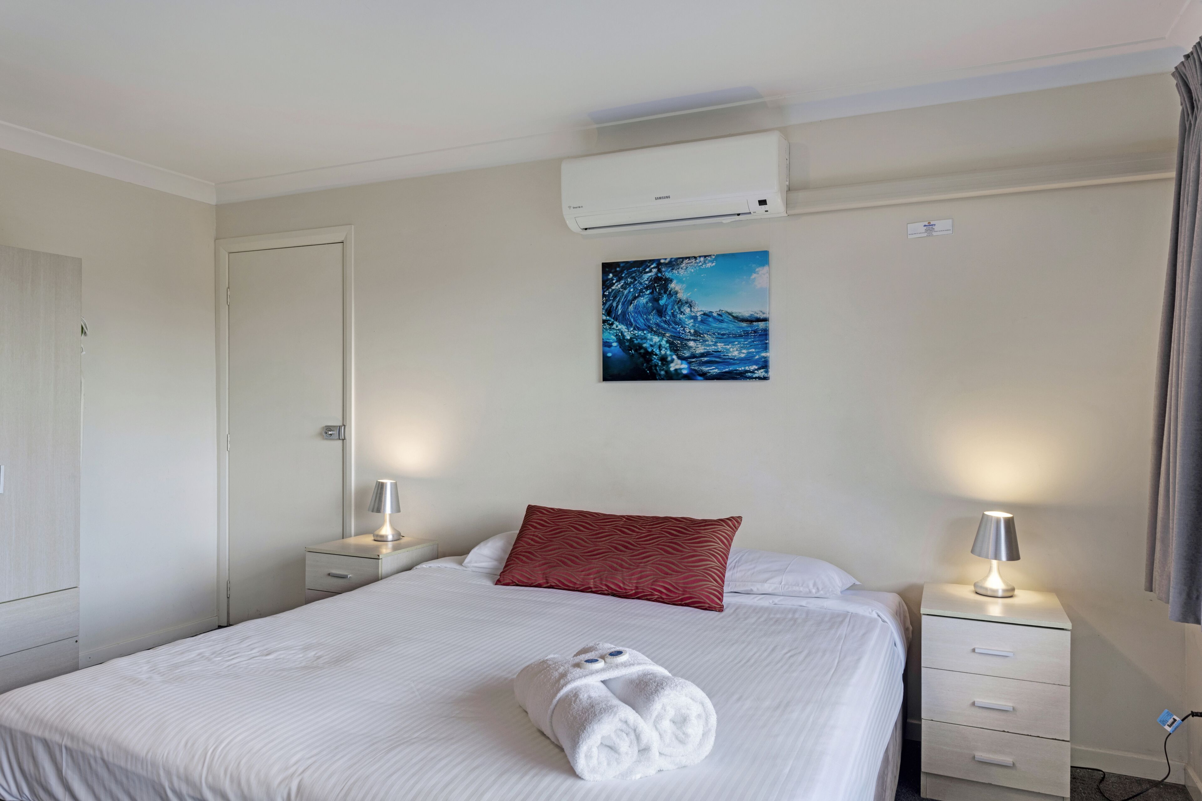 Una habitación de hotel con una cama, una almohada roja, una toalla blanca, una mesita de noche con una lámpara y un aire acondicionado en la pared.