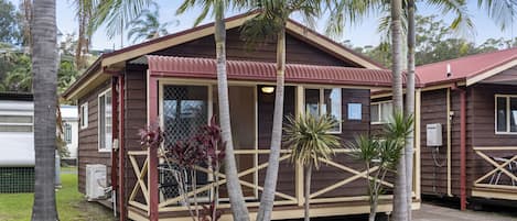 A small, single-story building with a brown exterior and a red roof. It has a porch with a railing and several palm trees in front.
