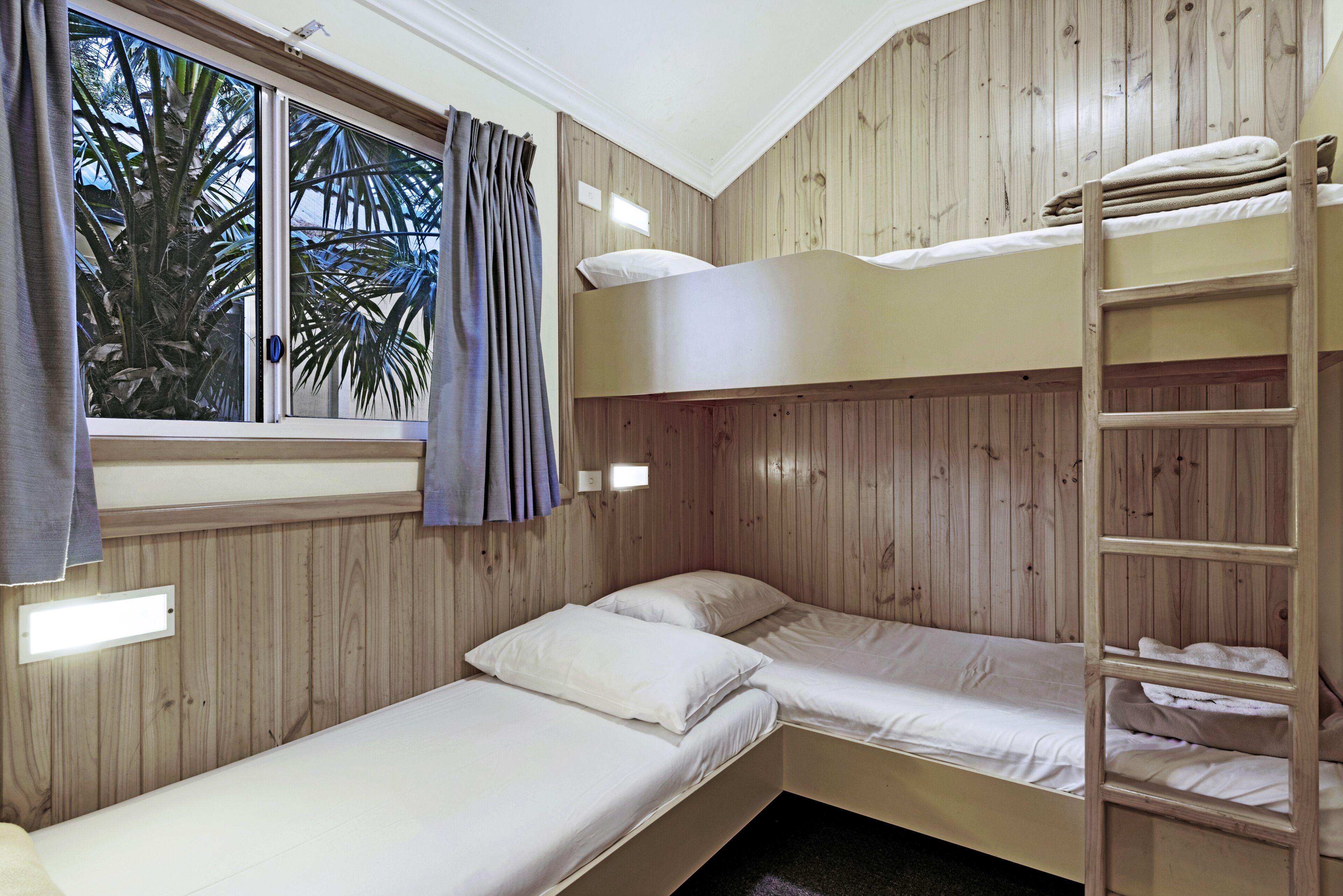 A bunk bed room with wooden paneling, a window with curtains, and a ladder.